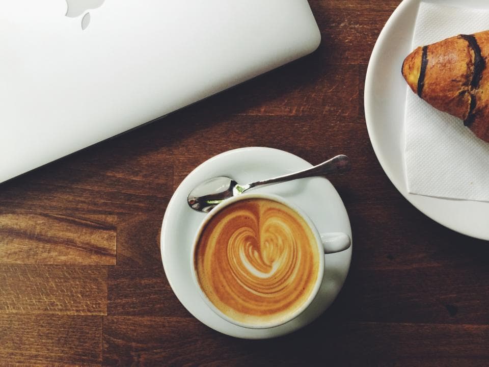 A flat white with leaf latte art beside a laptop and a croissant