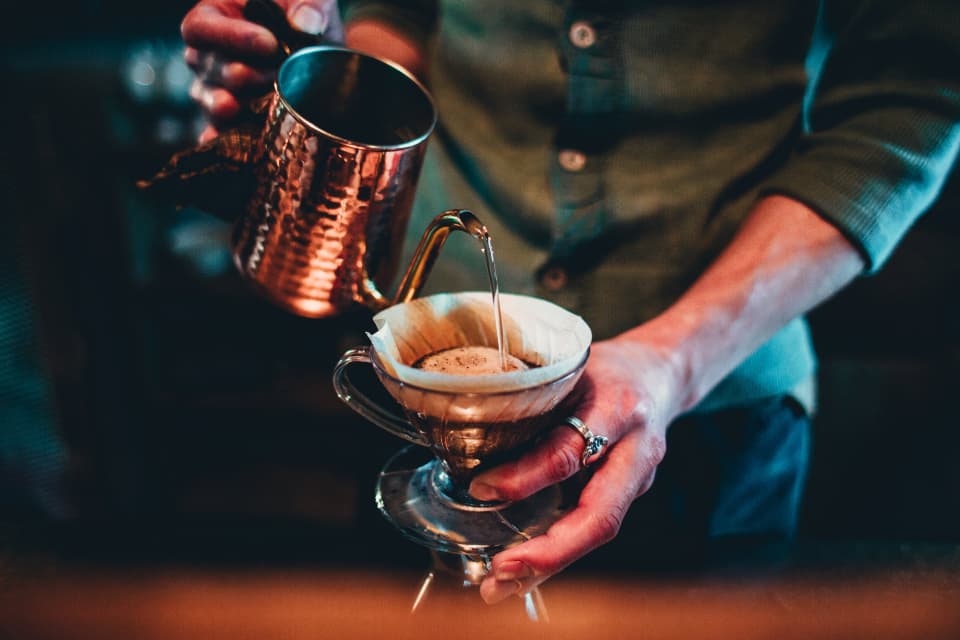 A barista pouring hot water from a copper kettle into a pour-over filter