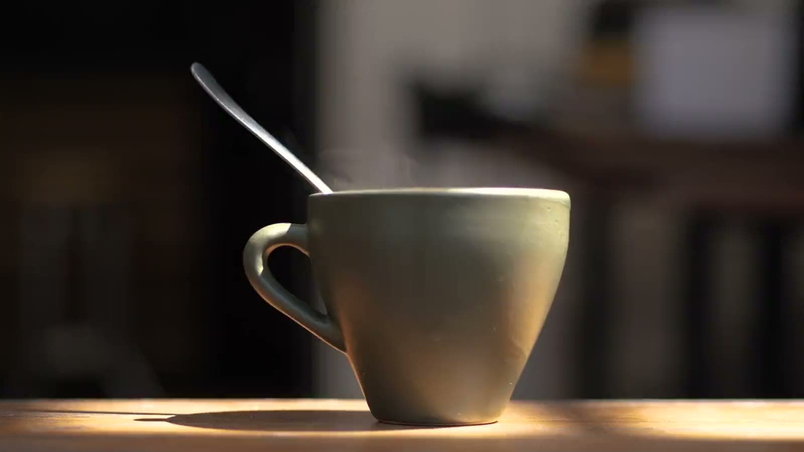 A steaming cup of coffee with a spoon on a sunlit wooden counter, the shop dark behind it